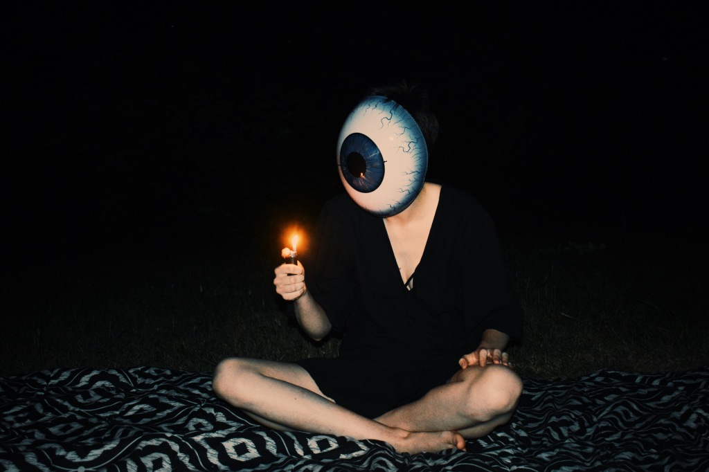 A figure sits cross-legged outdoors in the dark, wearing a mask that resembles a large eyeball and lighting a lighter.